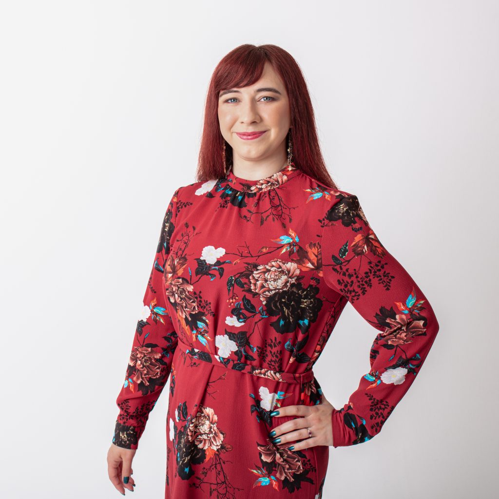 Female with dark red hair, wearing a red dress with flowers on. One hand is resting on hip. Plane white/grey background